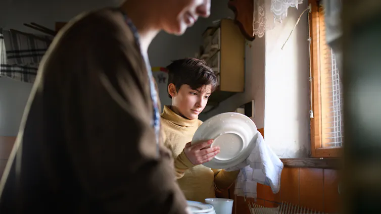 A young trans child washes dishes next to his mother.
