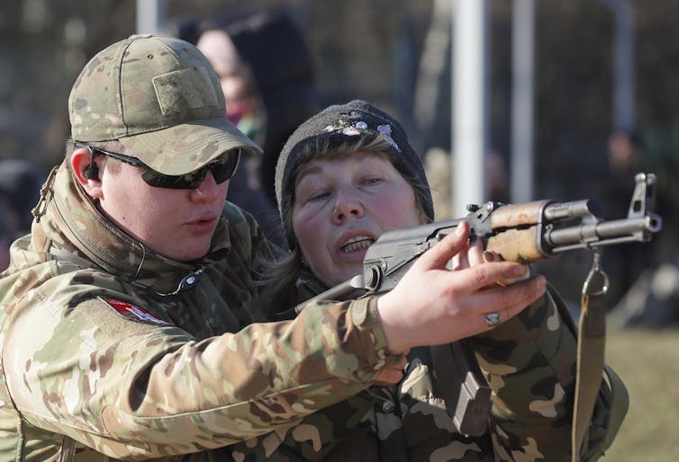Man teaching woman how to hold a rifle, both in army uniforms.