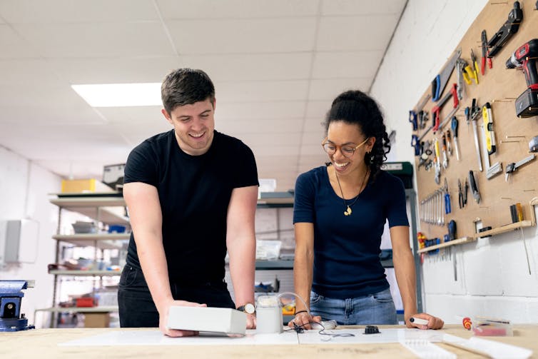 A man and a woman stand in front of a workbench with tools in the background.