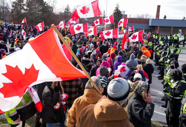 A line of armed police officers stand in front of a group of people waving Canadian flags.