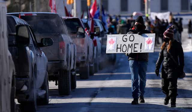 People holding a 'go home' sign walk past a line of trucks on the street.