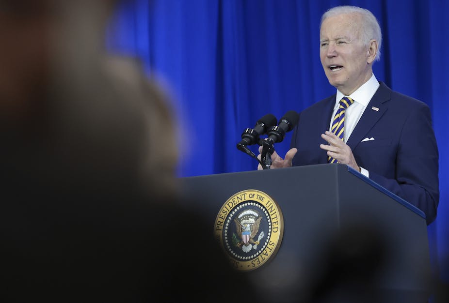 A white-haired man in a blue suit speaks into a microphone at a lectern emblazoned with the U.S. presidential seal.
