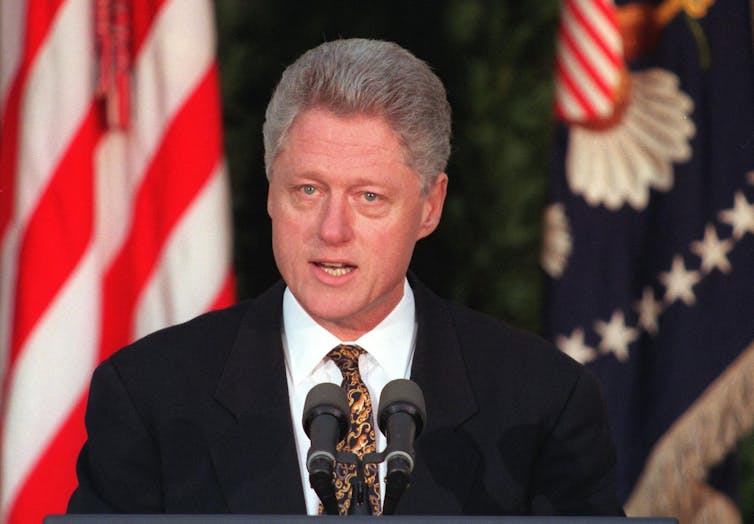 A man in a dark suit speaks into microphones, with flags in the background.