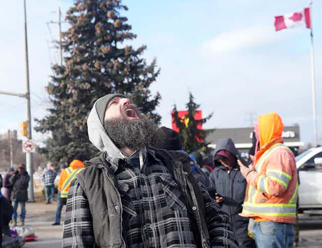A bearded white man yells towards the sky with Canadian flags behind him.