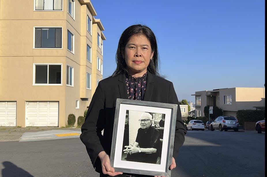 A woman standing in front of an apartment building holds a photograph of a man