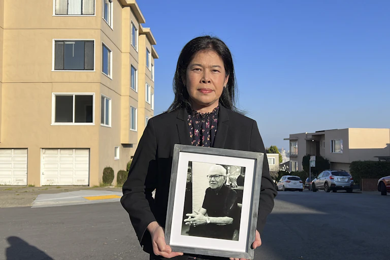 A woman standing in front of an apartment building holds a photograph of a man