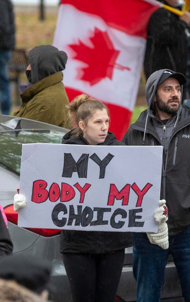 A woman carries a My Body, My Choice sign among other protesters. A Canadian flag is waved behind her.