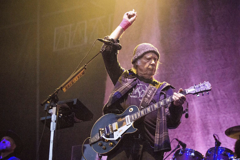 Guitarist seen on a stage against a purple backdrop with his hand in the air holding a guitar.