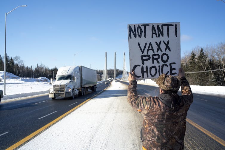 A man stands at a side of a highway holding a sign that says Not Anti-Vax, Pro-Choice