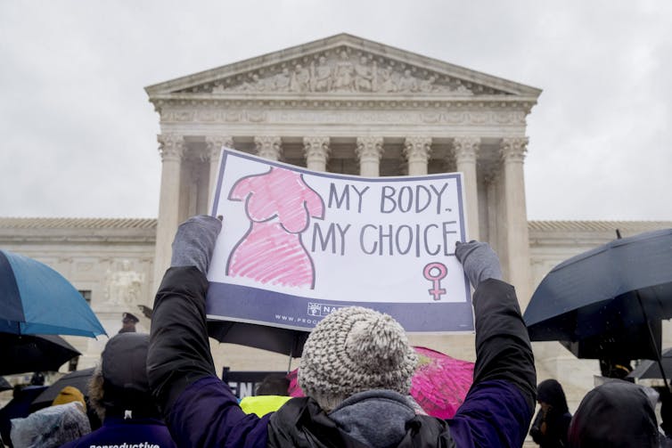 A person in a grey winter hat with a pompom holds up a sign that reads 'my body, my choice' in front of a court building with columns.