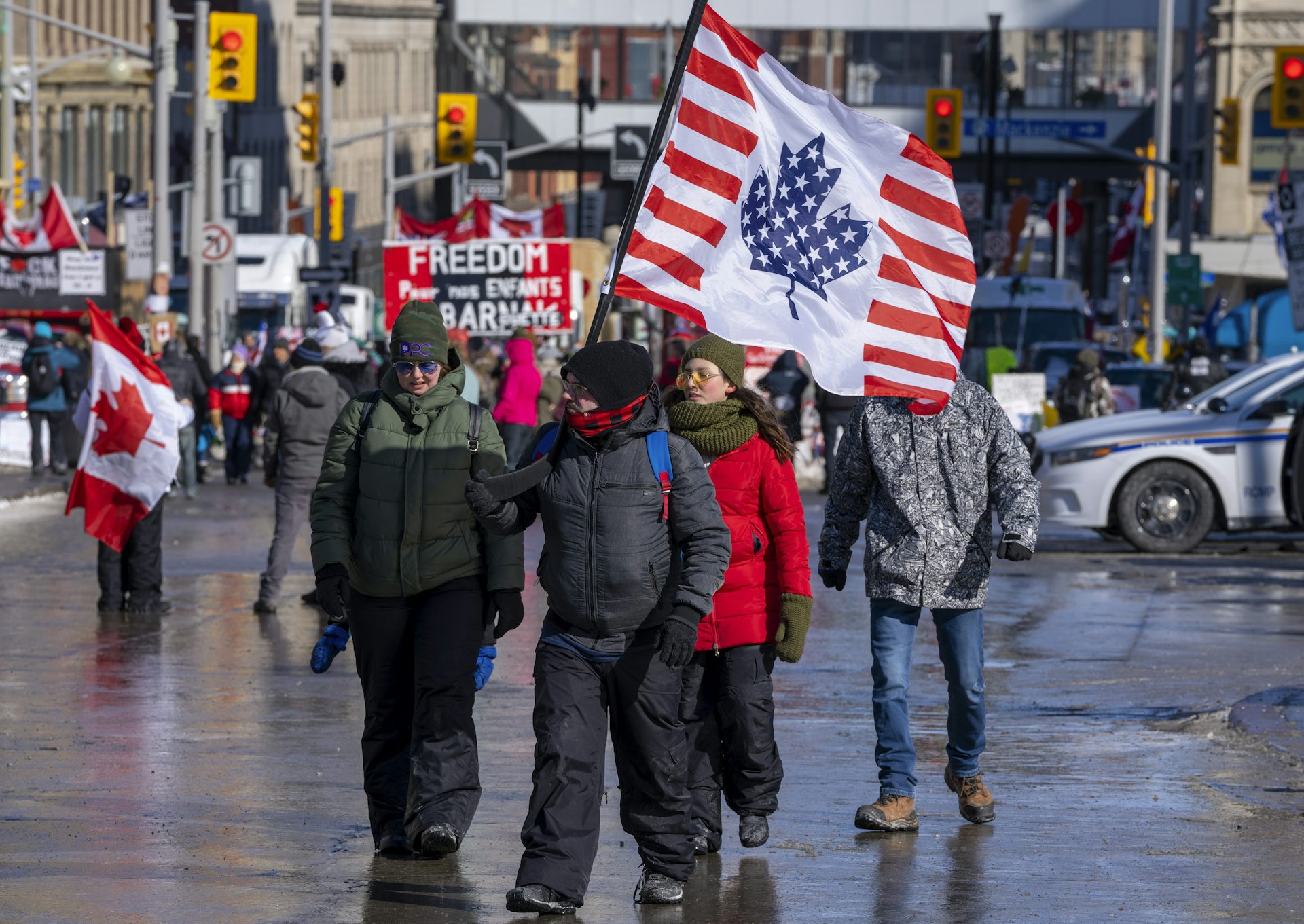 A group of people wearing winter gear walk down the street. They are holding a Canadian flag with the colouring of the U.S. flag.