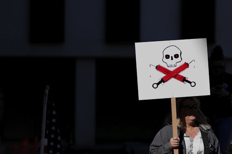 A person in shadow holding a sign depicting a skull and crossed vaccine syringes