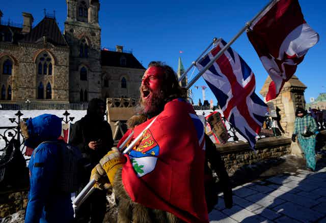 A man walks down the street in front of Parliament Hill, his face painted red as he carries the Union Jack and the Canada Flag