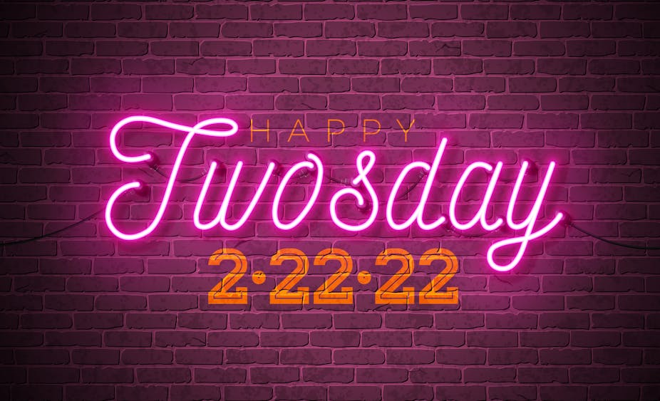 A neon sign against a brick wall reads, "Happy Twosday 2/22/22."