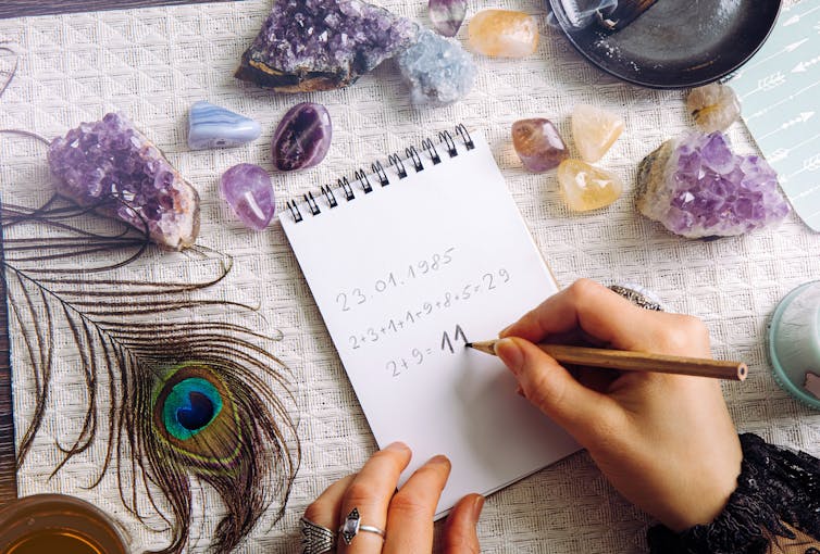 A photograph of someone's hand as they do numerology calculations at a table covered with geodes and a feather.