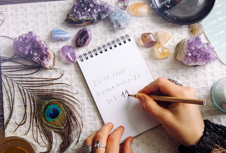 A photograph of someones hand as they do numerology calculations at a table covered with geodes and a feather