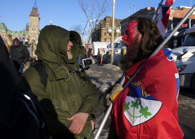 Um homem vestindo uma bandeira vermelha e pintura facial vermelha grita com outro homem vestindo um casaco verde-oliva