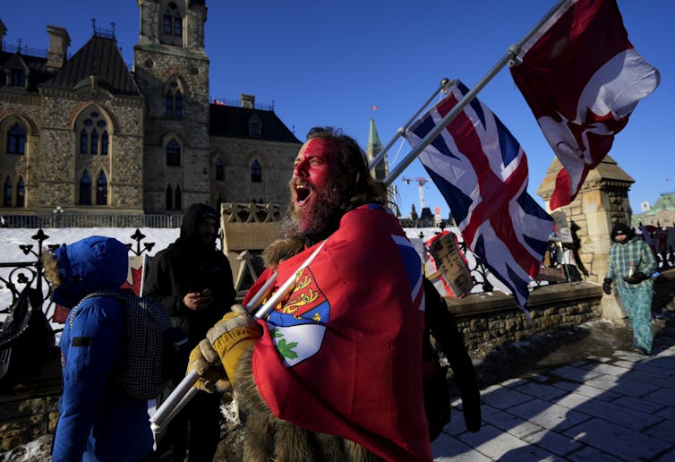 A man in red face paint shouts while carrying a flag in front of Parliament.