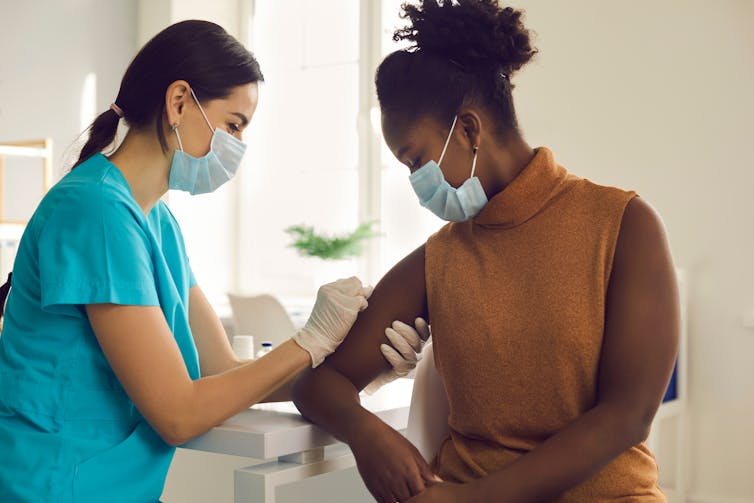 A masked nurse vaccinates a young African-Australian woman.