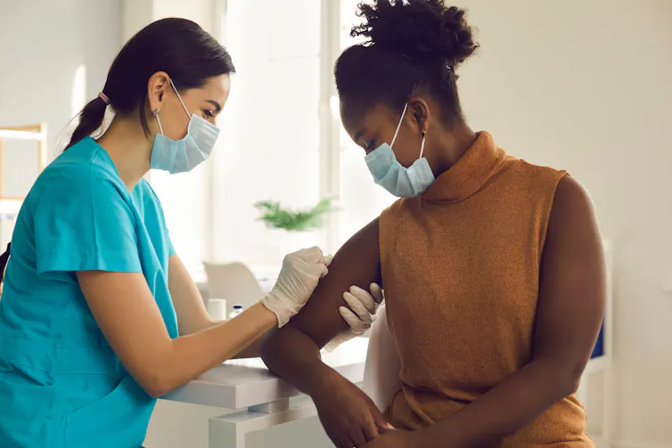 A masked nurse vaccinates a young African-Australian woman.