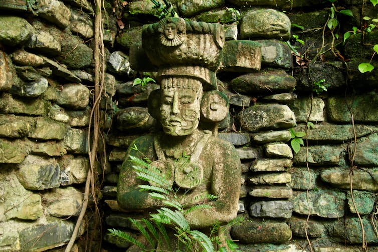 A fern growing in front of a stone human figure with a rock-pile wall behind.