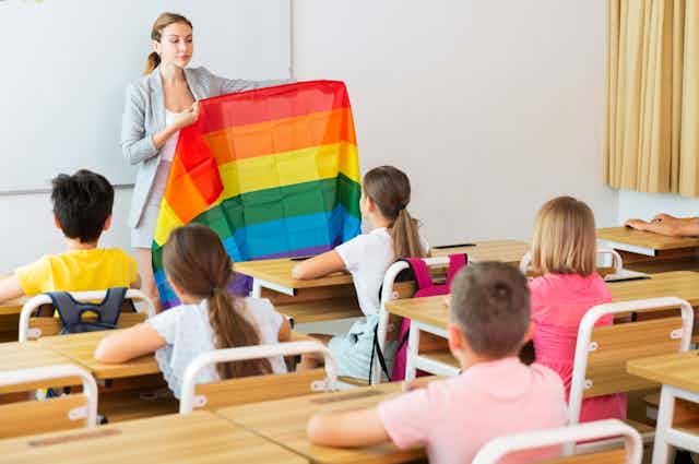 A teacher holds a rainbow flag at the front of a classroom of young students