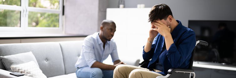 Two men are sitting in a living room, with one holding his head in his hands.