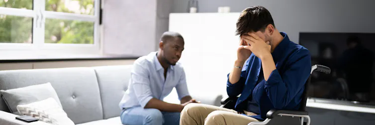 Two men are sitting in a living room, with one holding his head in his hands.