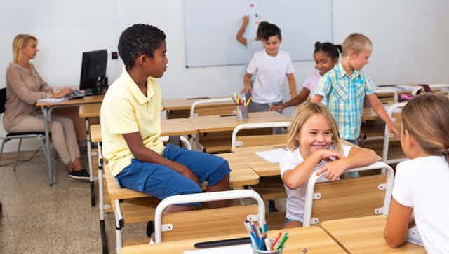 Enfants en train de parler dans une salle de classe. Leur prof est devant un ordinateur.