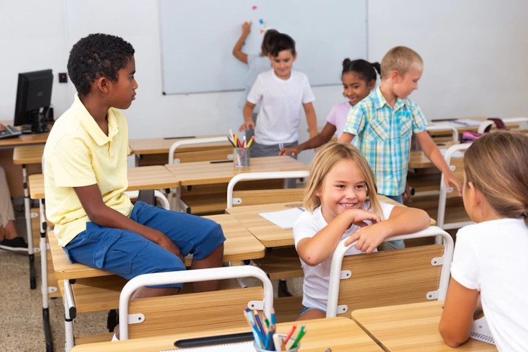 Enfants en train de parler dans une salle de classe. Leur prof est devant un ordinateur.