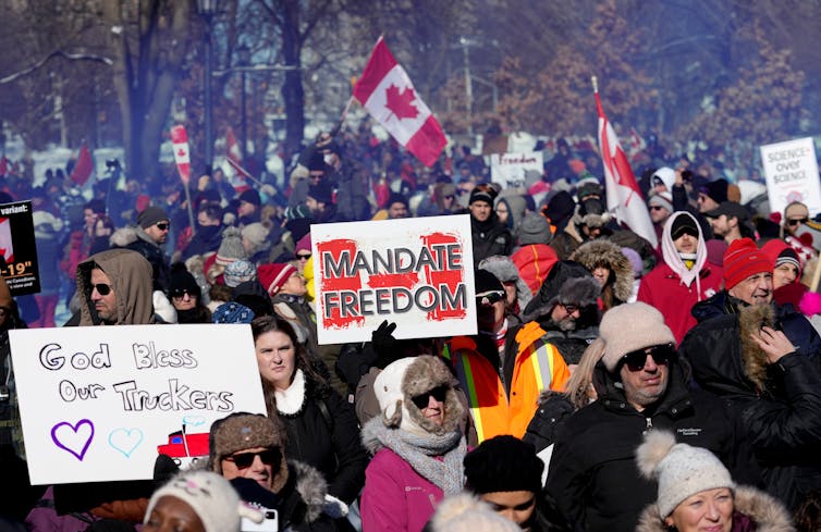 A crowd of people holding signs. One visible sign says 'MANDATE FREEDOM' and another says 'God Bless Our Truckers.'