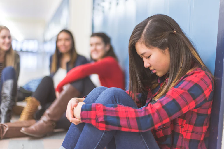 A young woman sits alone with her head down while other young women in the backgrond laugh at her