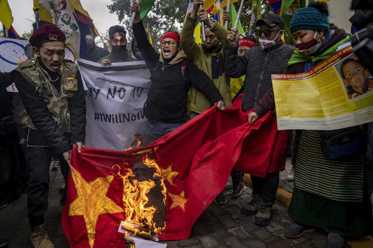 A group of people holding protest signs stand around a flag that's on fire.