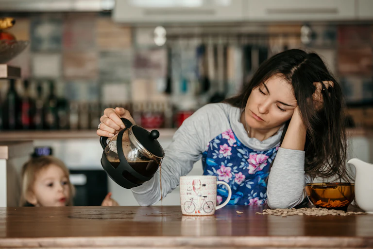 A woman with her eyes closed supports her head with one arm and pours coffee onto the table, missing her mug, with the other. A child watches in the background.