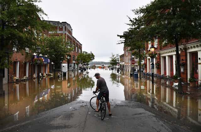 A man on a bicycle looks at flood water covering most of a downtown street. 