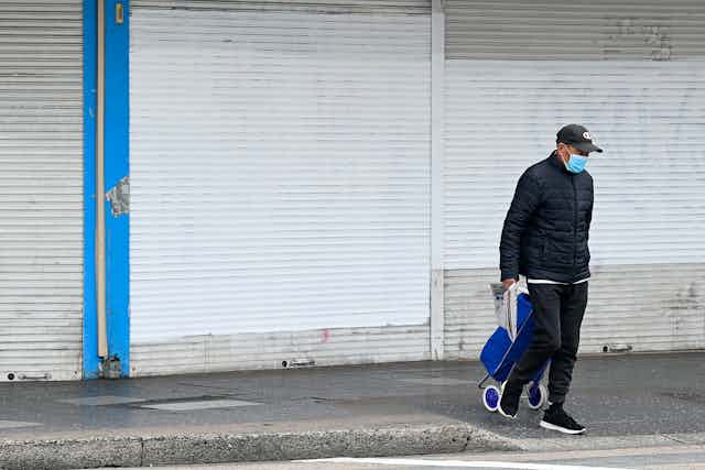 A man walks while wearing a mask and wheeling a shopping trolley