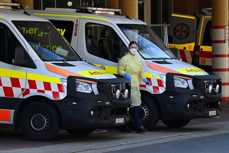 Ambulances line up at a hospital, while a healthcare worker in PPE stands outside.