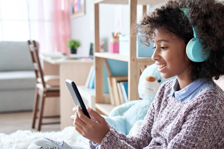 Girl wearing headphones looking at at her phone as she sits on her bed