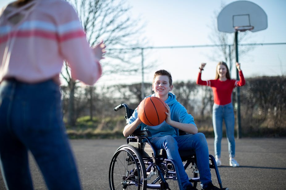 A young boy in a wheelchair passes a basketball to a young girl, while another girl cheers them on.