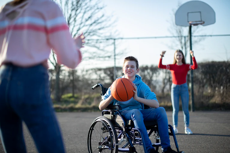 A young boy in a wheelchair passes a basketball to a young girl, while another girl cheers them on.