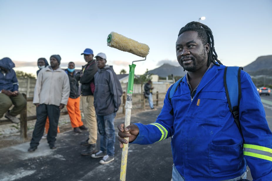 A dreadlocked man wearing an overall holds a paint roller on the side of a road in the hope of finding a casual painting job. Five other men, also seeking work, look on behind him.