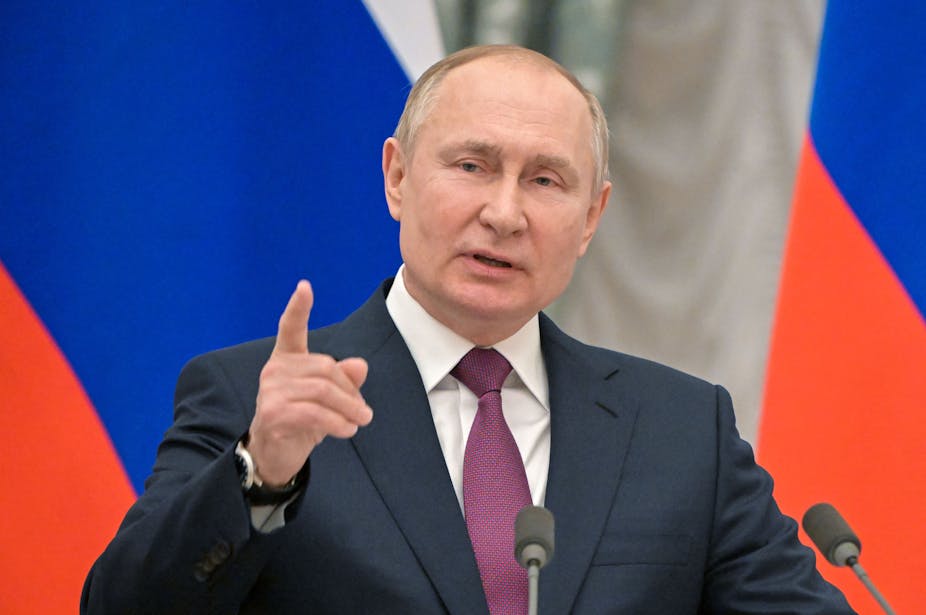 Vladimir Putin in a suit in front of Russian flags points a finger towards the camera.