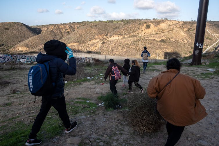 People wearing backpacks are shown walking in a desert, with a graffiti-covered fence ahead of them.