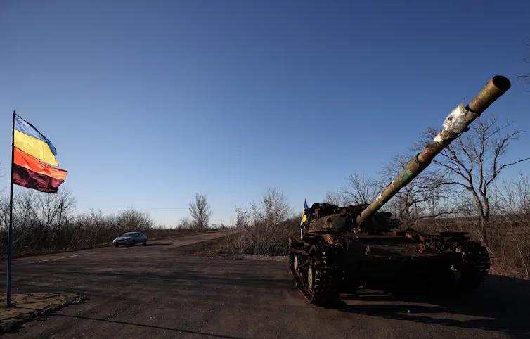 A tank sits by the side of an open road with a flag visible.
