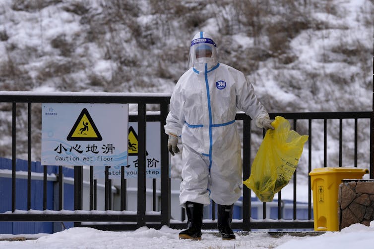 A person in a protective suit and face shield holding a garbage bag