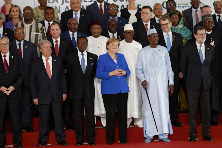 A group of people stand on a red carpet in front of signage; some wearing suits, others in robes