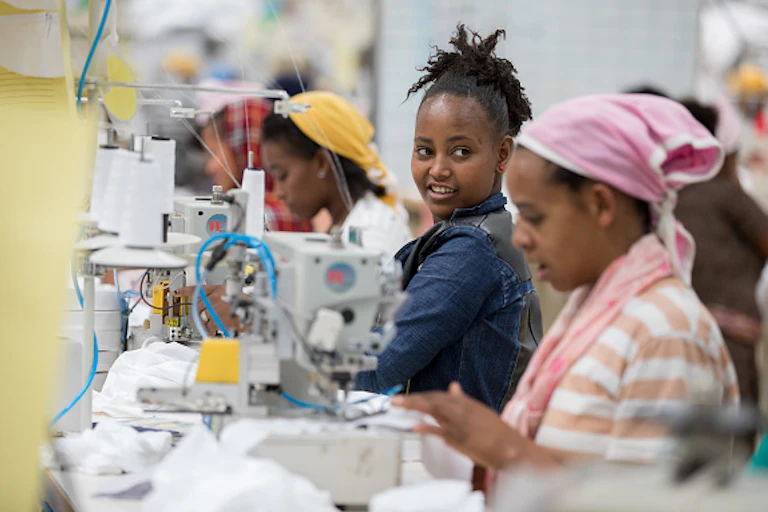 Women sit in a line at sewing machines, one woman turning to look at her co-worker.