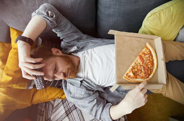 Un hombre con aspecto depresivo tumbado con un mando a distancia y una pizzza.