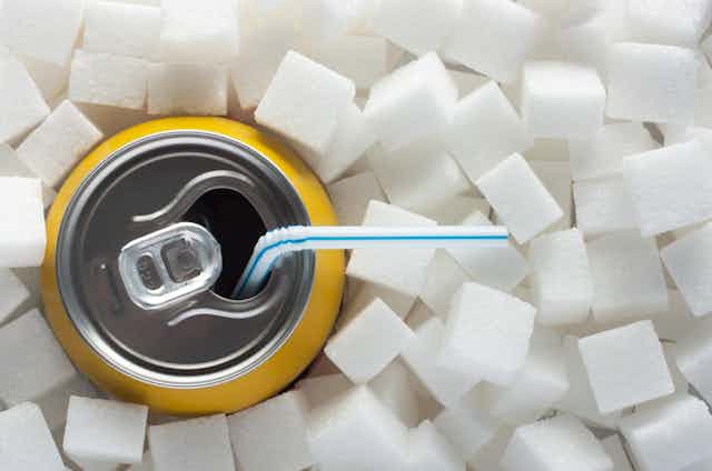 A can of fizzy juice with a straw sitting in a big heap of sugar lumps.