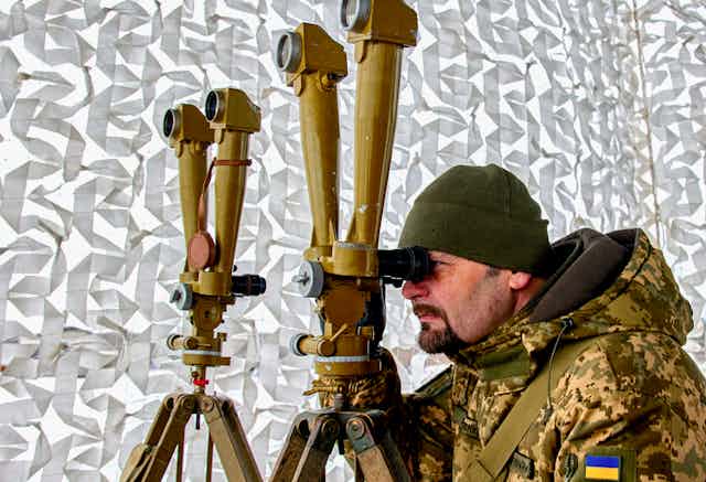 A man wearing a Ukranian military uniform looks through a specialised long-distance viewing apparatus during a training exercise.